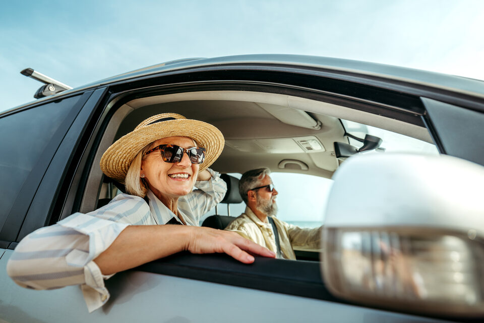 A snowbird couple driving south, the passenger leaning out of the window in the sun